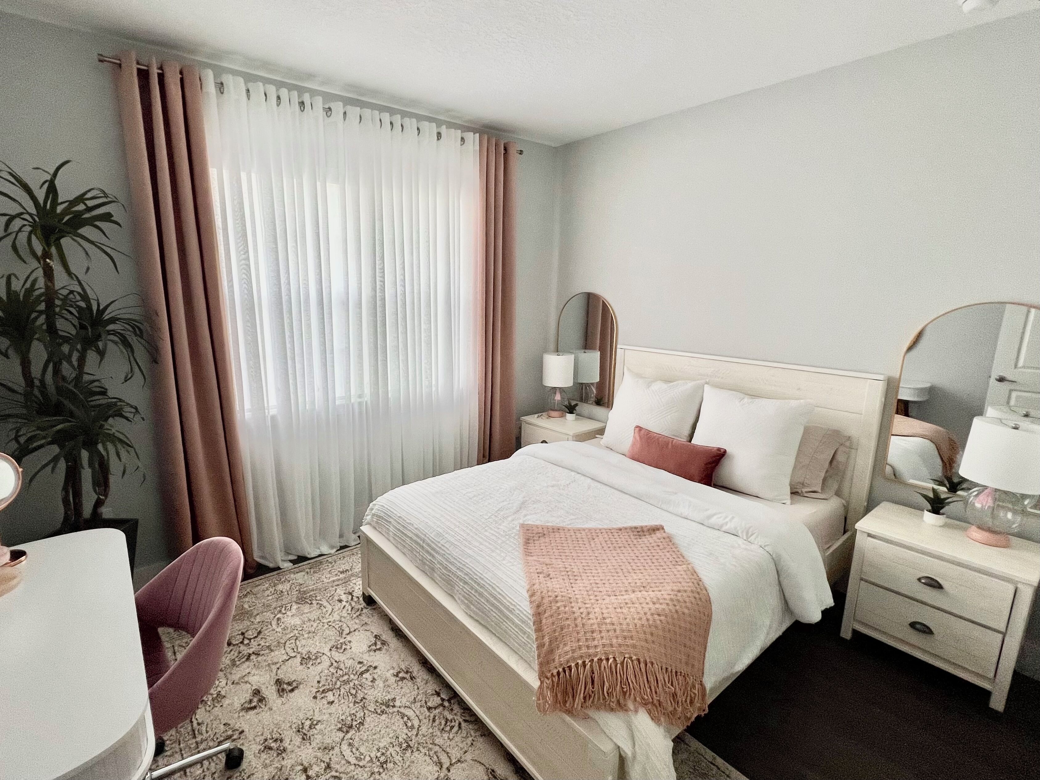 A hotel room with a bed, bedside tables, a chair, a desk, and a large window with curtains.