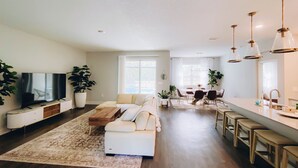 A modern living room with a flat-screen TV, a sofa, a coffee table, and a dining area with a table and chairs.