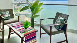 Un balcon avec des fauteuils en osier, une table colorée à rayures et une plante en pot.