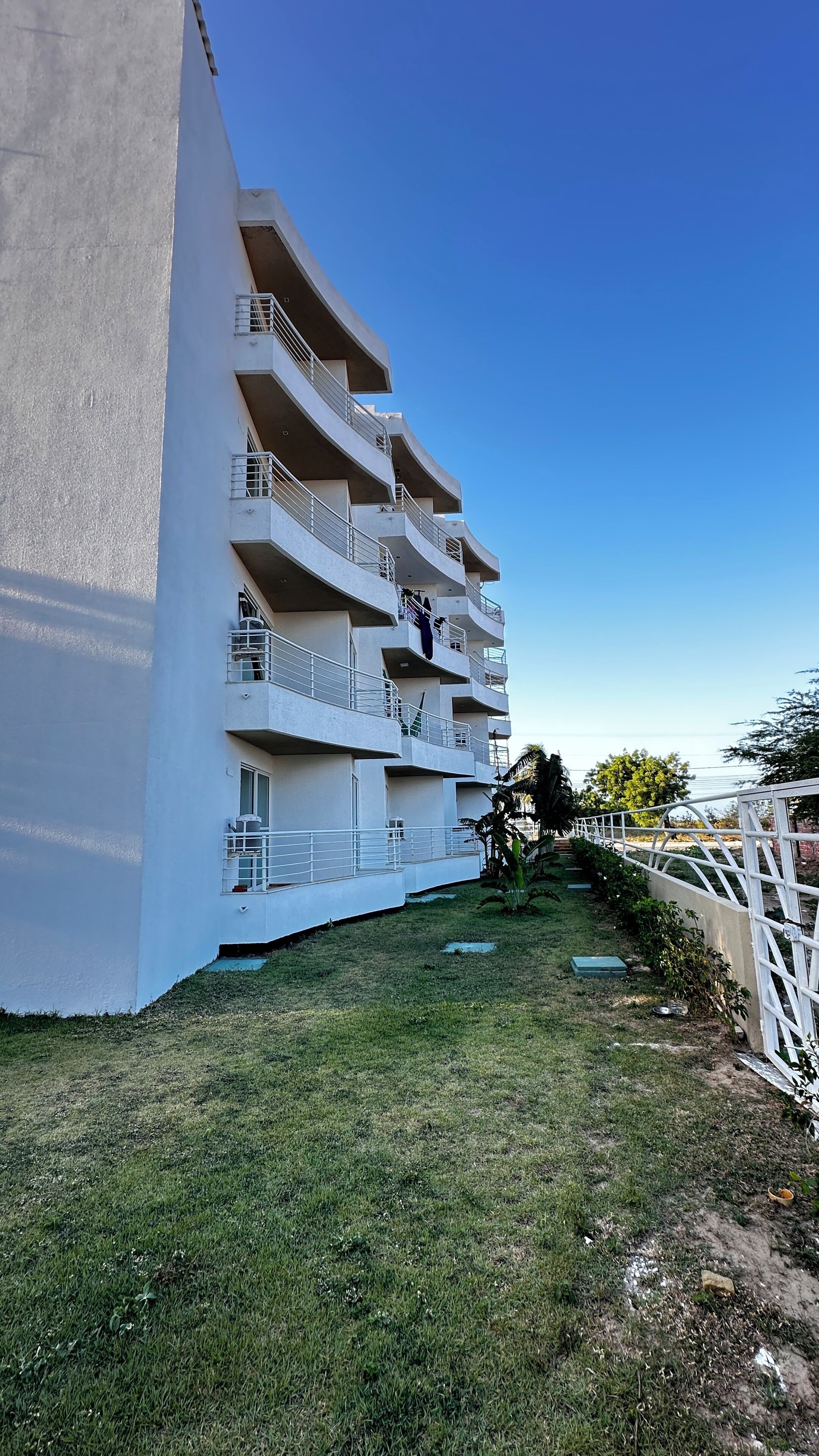 Un immeuble à plusieurs étages doté de balcons, avec un espace verdoyant devant et un ciel bleu dégagé.