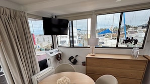 A room with a round wooden table, a white chair, a wooden dresser, a TV mounted on the wall, and a view of boats through large windows.