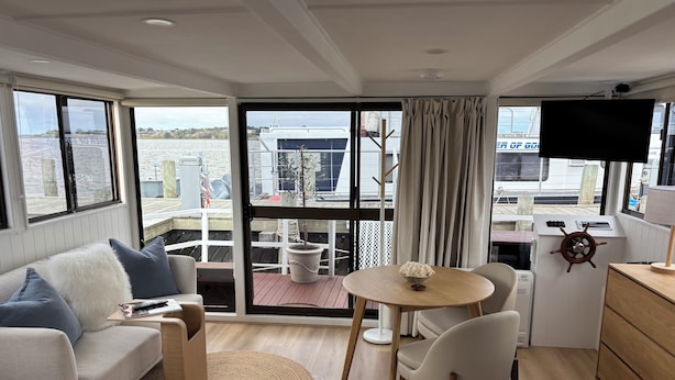 A modern cabin with a bed, a sofa, a dining area with a table and chairs, a TV, and a view of the water and dock outside.