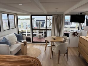 A modern cabin with a bed, a sofa, a dining area with a table and chairs, a TV, and a view of the water and dock outside. - Piccolina Floating Accomodation (Goolwa)