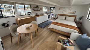 A compact, modern room with a bed, a small dining table, a kitchenette, and a seating area with a magazine.