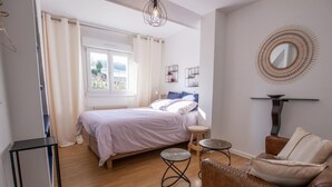 Un dormitorio con cama, mesita de noche, una silla, una pequeña mesa redonda, un espejo y una ventana con cortinas.