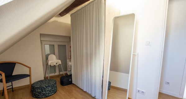 A small, well-lit room with a sloped ceiling, a wooden chair, a round ottoman, a white curtain, and a mirror. - A la Croisée des Louvières (LE MESNIL SUR OGER)