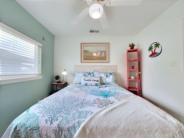A bedroom with a bed, a ceiling fan, a nightstand with a lamp, a framed picture on the wall, a shelf with decorative items, and a window with blinds.
