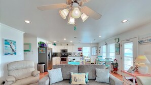 A spacious living room with a grey sofa, a ceiling fan, and a kitchen in the background.