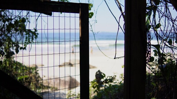 A beach view through a wire fence with greenery on the sides. - Solitude Cabin - Sailor (Pântano do Sul)