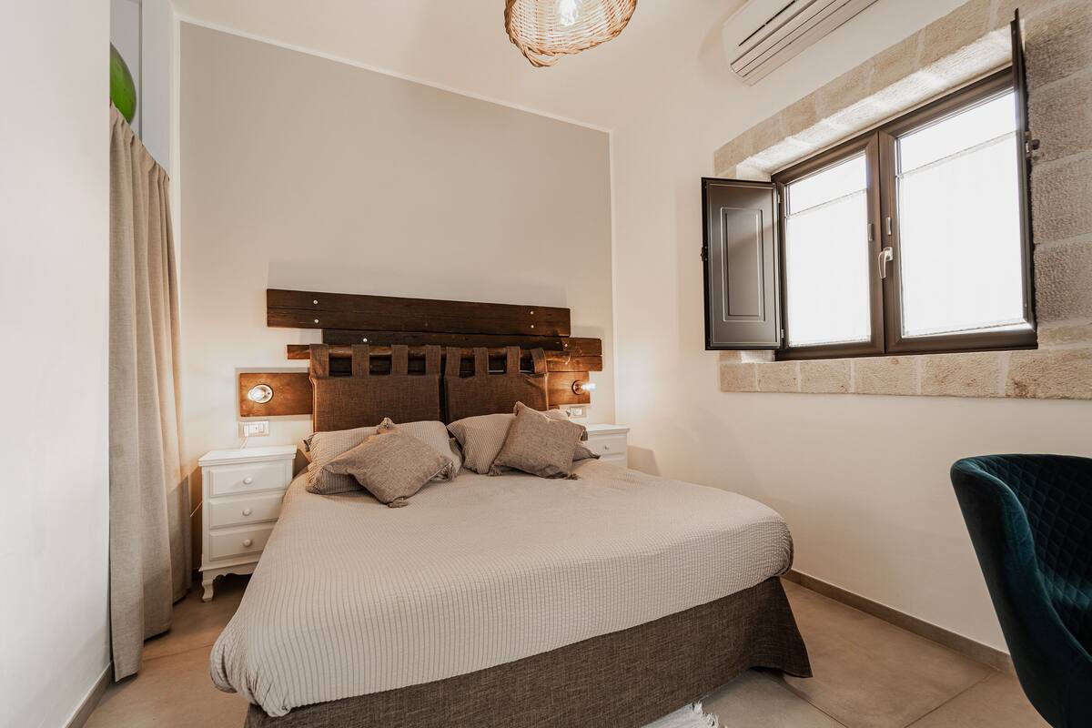 Un dormitorio con cama, mesitas de noche, una silla y una ventana con cortinas.