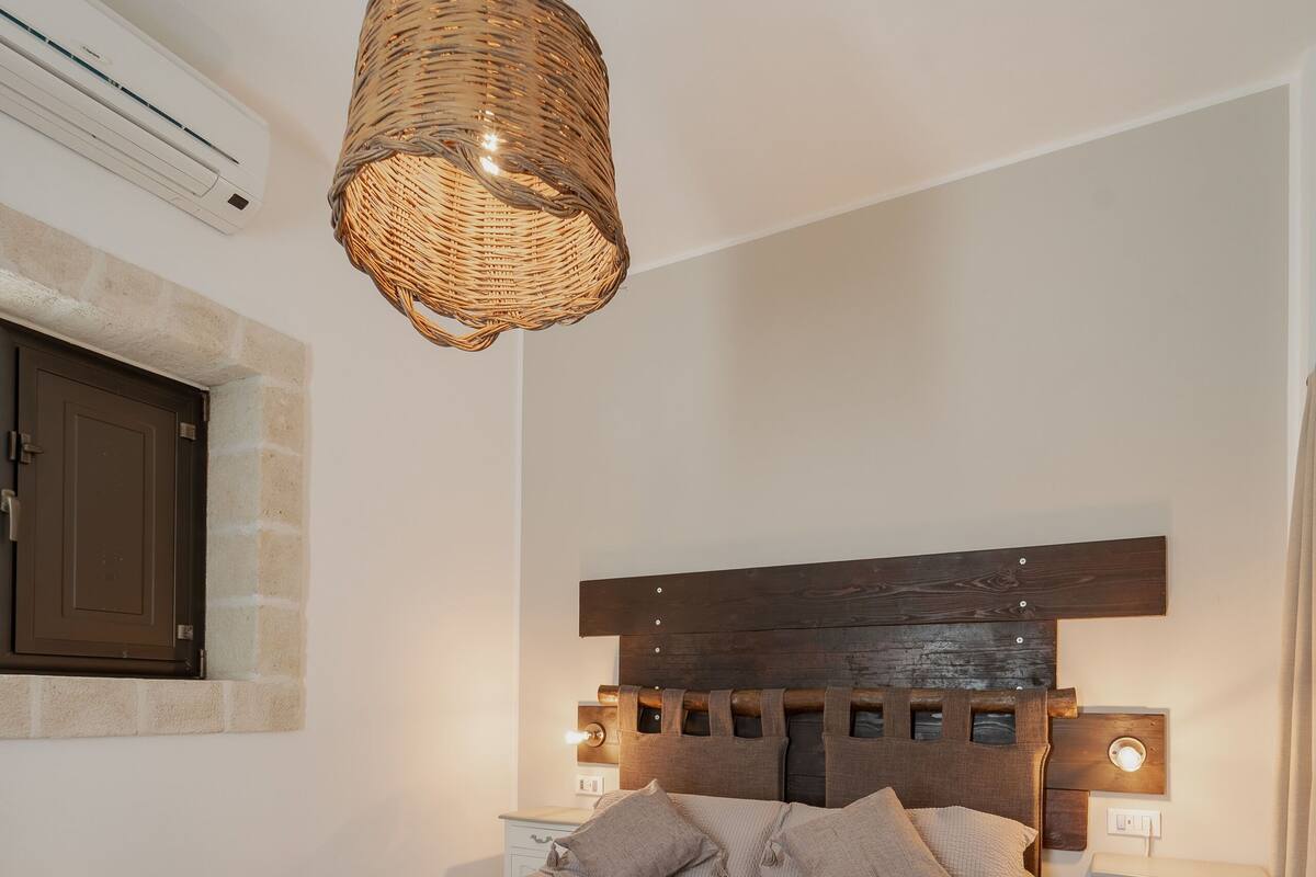 Un dormitorio con cabecera de madera, mesita de noche, lámpara colgante de mimbre y mesita auxiliar.