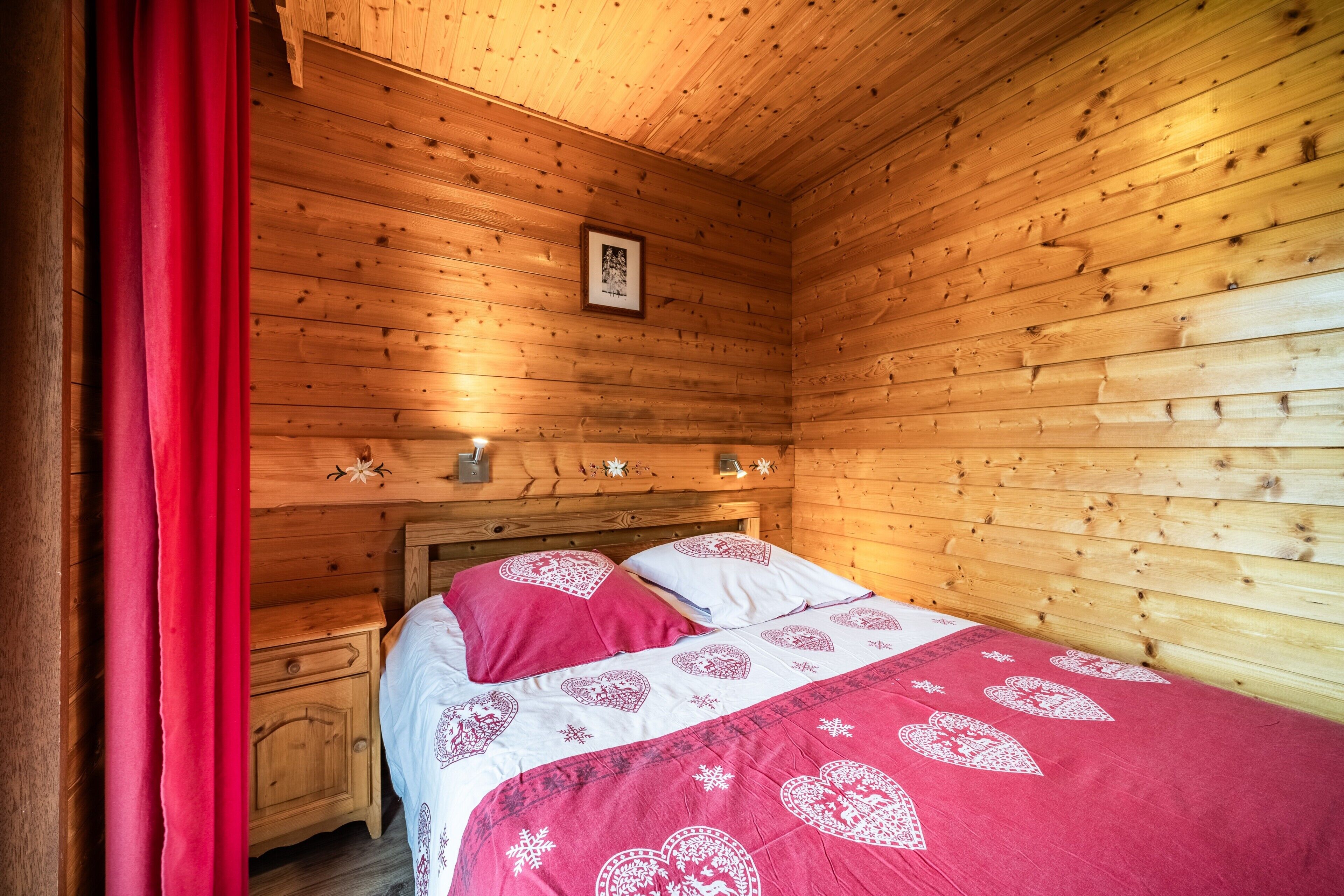 Een houten slaapkamer met een bed, een nachtkastje, een ingelijst schilderij aan de muur en een rood gordijn.