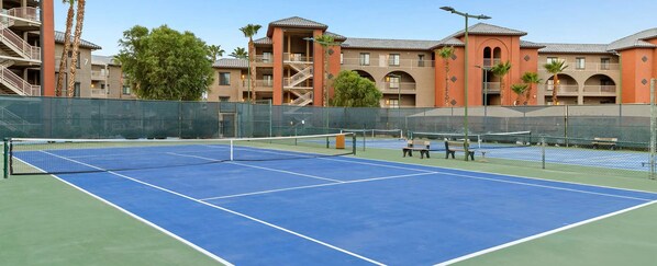 A tennis court with a net, surrounded by a fence, and adjacent to a building with multiple balconies and palm trees. - 2-bedroom resort in superb Indio with fitness room, AC. Enjoy relaxation (Indio)