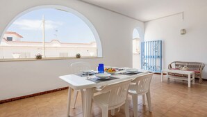 Comedor con una mesa blanca preparada para cuatro comensales, sillas blancas y vistas a través de amplias ventanas arqueadas.