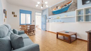 Una sala de estar con un sofá, una mesa de comedor con sillas, una mesa de centro de madera, un ventilador de techo, un televisor montado en la pared y un cuadro grande de un barco.