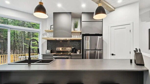 A modern kitchen with a large island, stainless steel appliances, pendant lighting, and a view of the forest. - Le Ô Calm |Spa| Hamac (Chertsey)