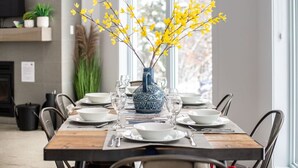 A dining table set with white plates, bowls, and cutlery, a blue vase with yellow flowers, and a window with a view of trees. - Le Ô Calm |Spa| Hamac (Chertsey)