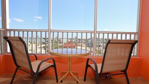 A balcony with two chairs and a round table, offering a view of a coastal area with buildings and palm trees.