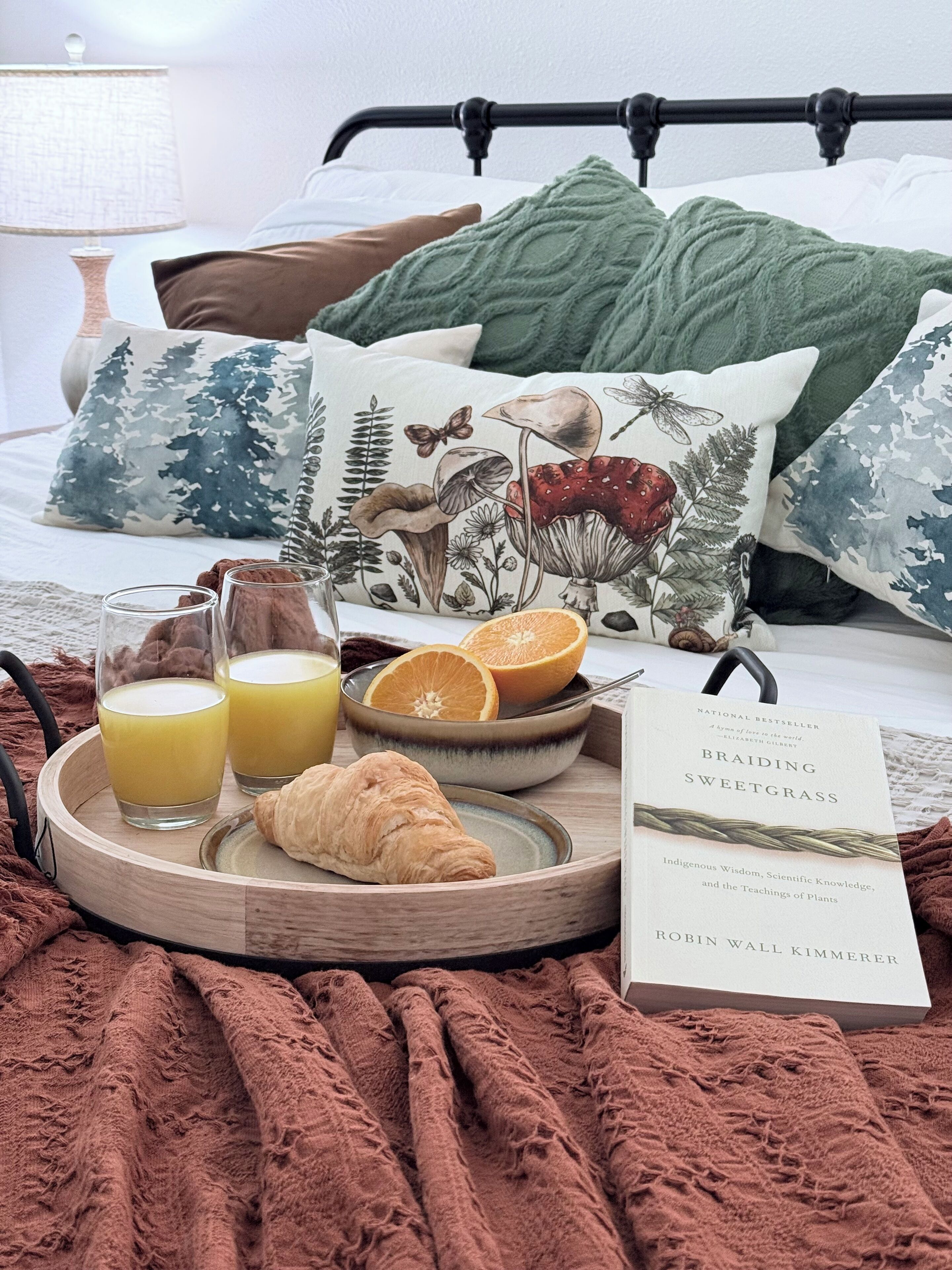 A neatly arranged breakfast tray with juice glasses, a croissant, and orange slices on a bed with a book titled "Braiding Sweetgrass" beside it.