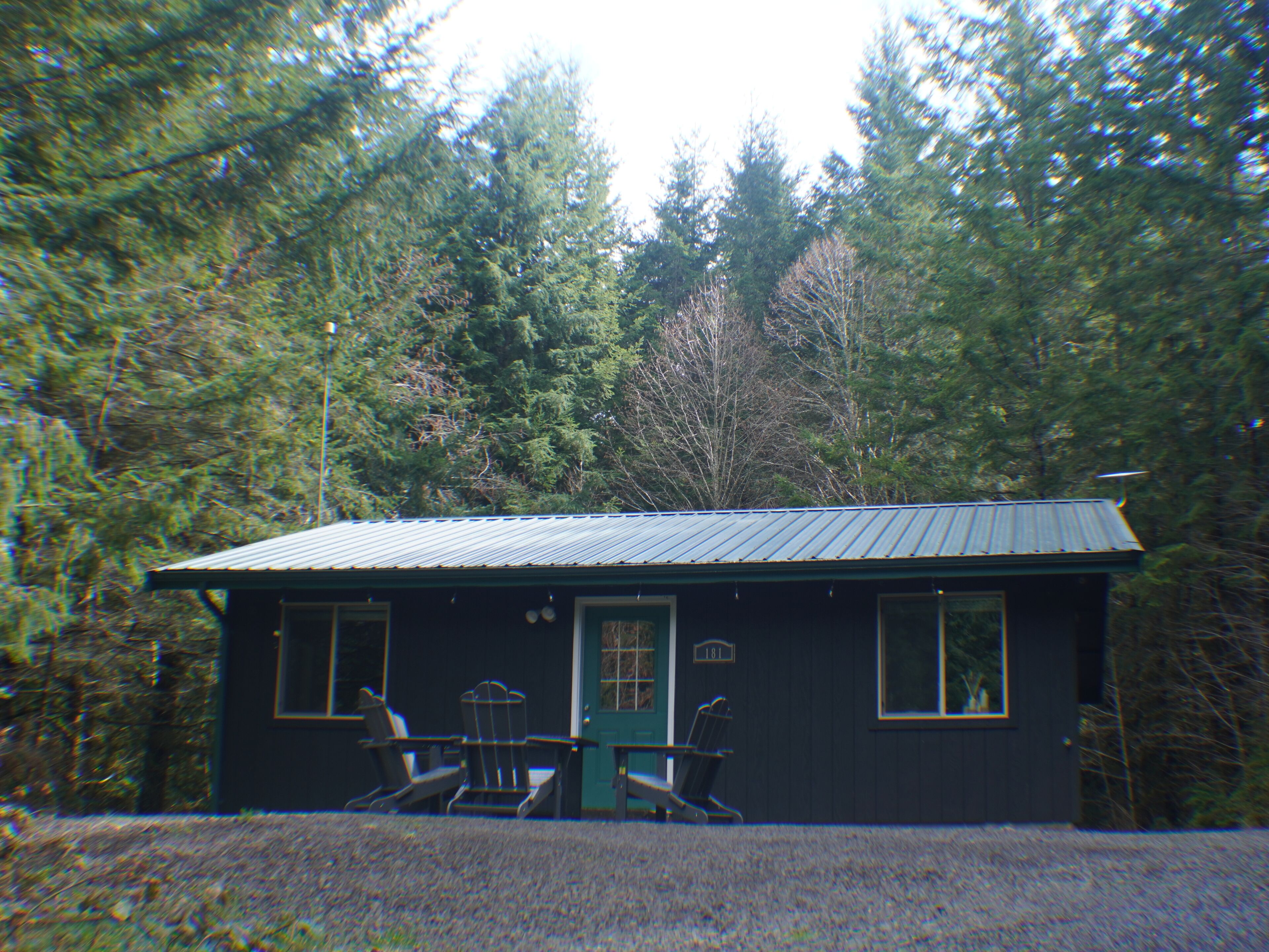 A cabin with a metal roof, two windows, and a door. There are two Adirondack chairs outside.