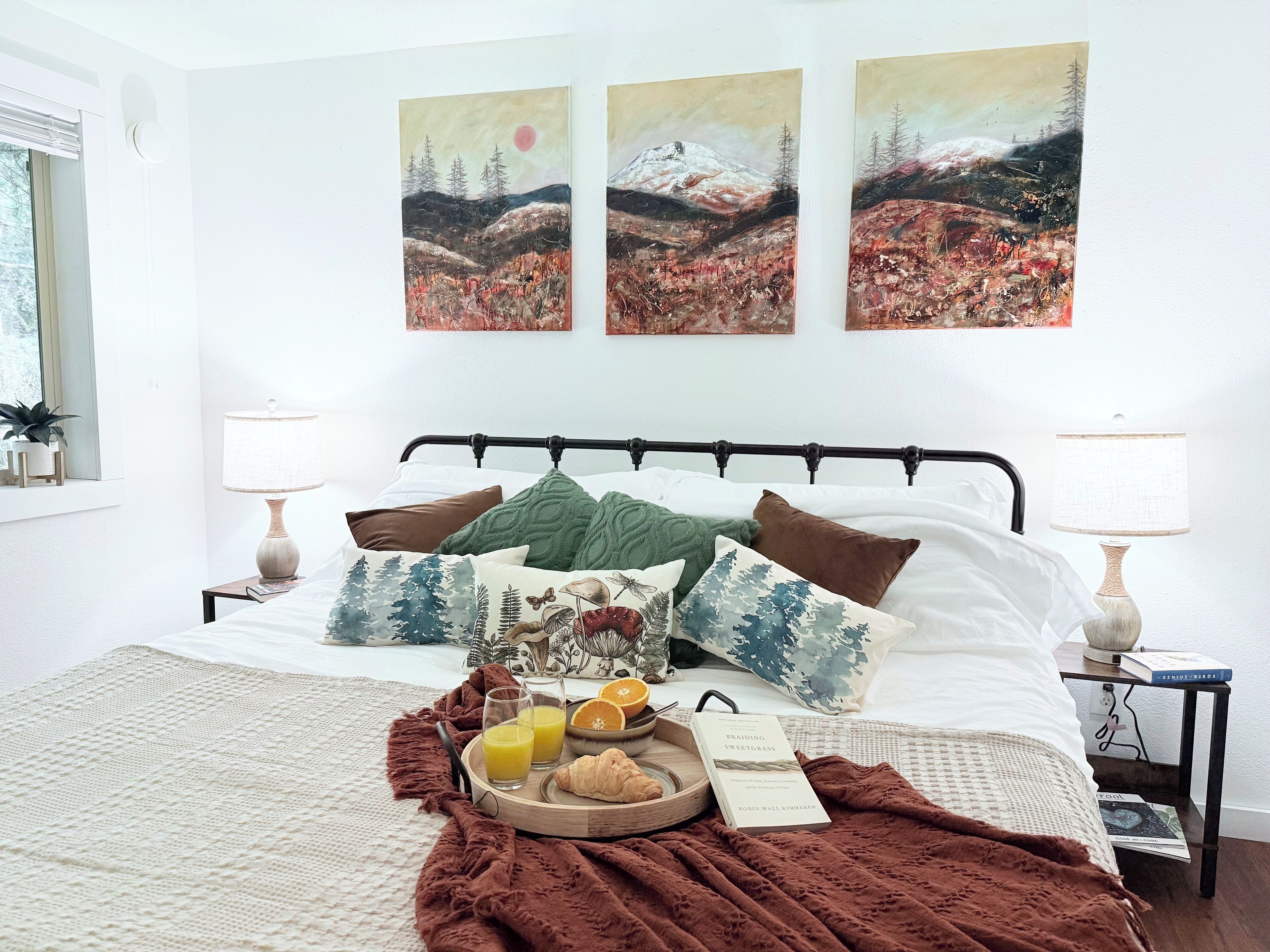 A neatly made bed with a variety of pillows and a blanket, a bedside table with a lamp and a book, and a tray with breakfast items.