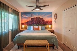 Ein Schlafzimmer mit Bett, Deckenventilator, Wandgemälde, Nachttisch mit Lampen, einer Bank und einer Tür.