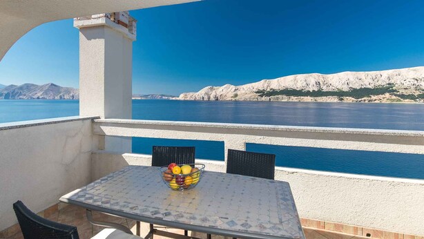 Ein Balkon mit Tisch und Stühlen, der auf ein Gewässer und hügeliges, felsiges Gelände blickt.