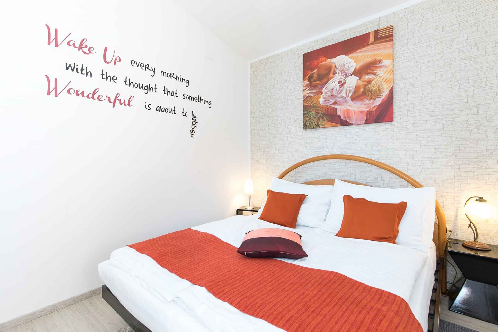 Ein ordentlich bezogenes Bett mit einer roten Decke und zwei orangenen Kissen, eine weiße Ziegelwand und ein motivierendes Zitat an der Wand.