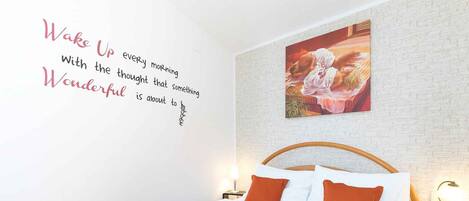Ein ordentlich bezogenes Bett mit einer roten Decke und zwei orangenen Kissen, eine weiße Ziegelwand und ein motivierendes Zitat an der Wand.