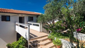 Ein weißes Gebäude mit rotem Ziegeldach, einer Treppe, die zu einem Balkon führt, und einem Garten mit verschiedenen Pflanzen und Blumen.