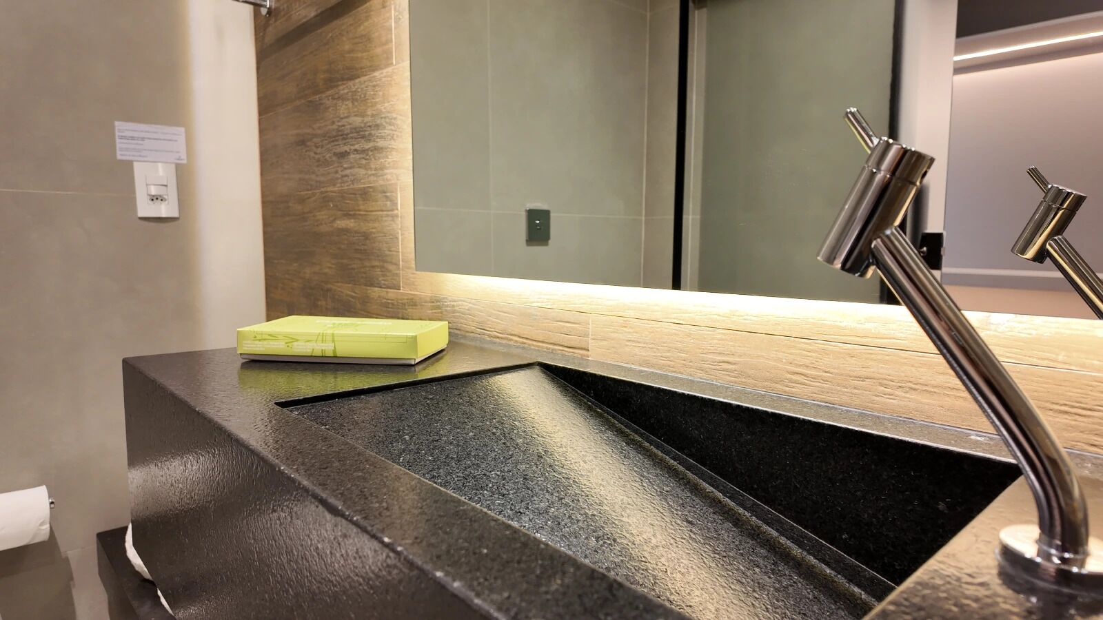 Un baño moderno con un lavamanos negro, un dispensador de jabón amarillo y un espejo con un panel posterior de madera.
