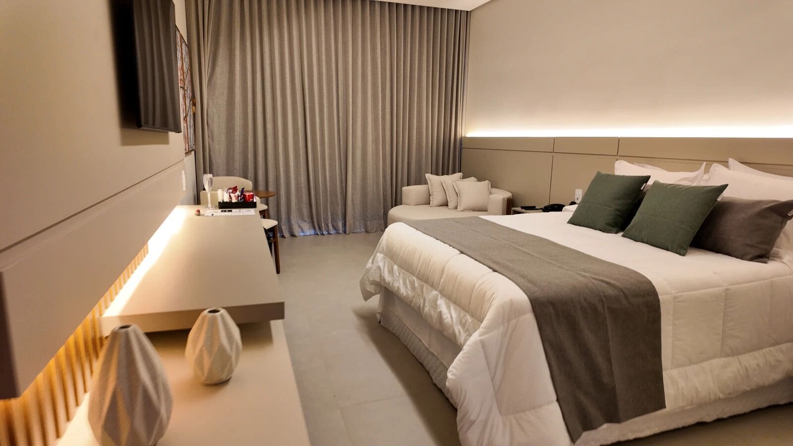 A modern hotel room with a neatly made bed, a bedside table with a lamp, and a large window with curtains.