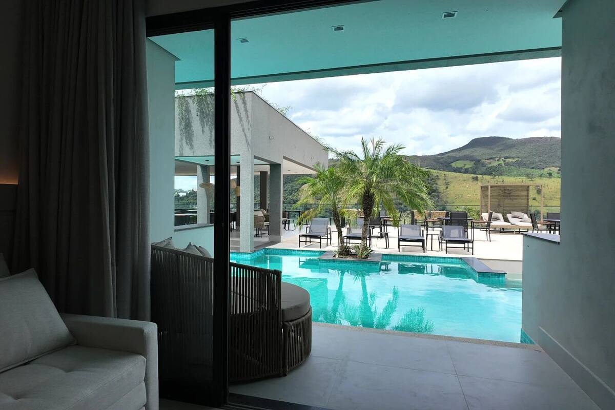 Una habitación de hotel moderna con un amplio ventanal corredera que da a un área de piscina con sillones reclinables y una palmera.