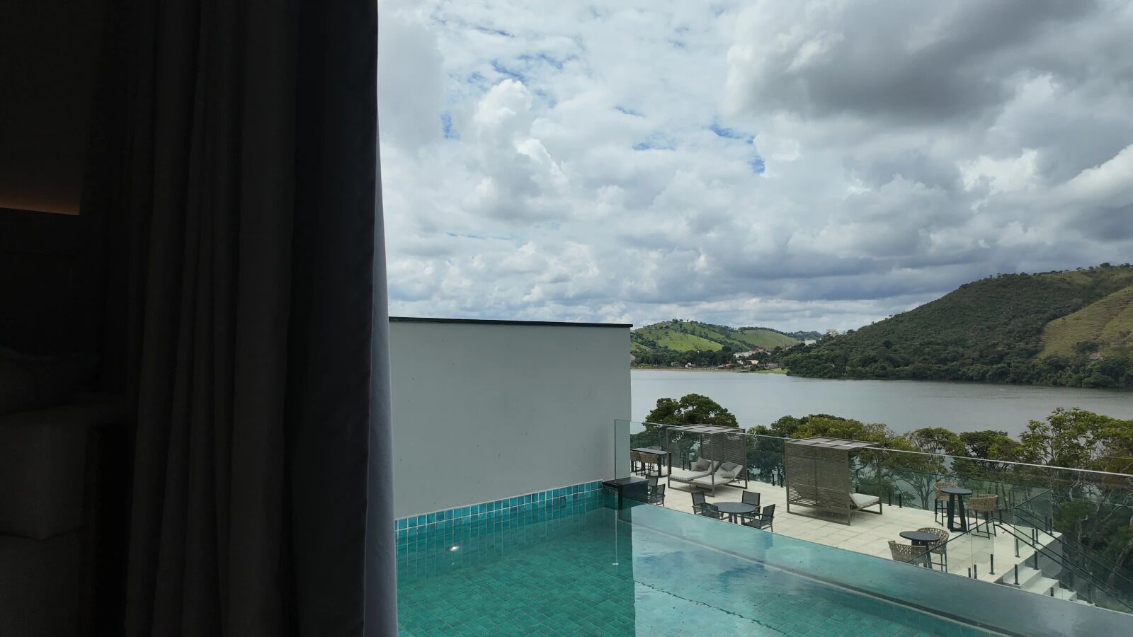 Een balkon met een zwembad met glazen rand, buitenmeubilair en uitzicht op een meer en heuvels.