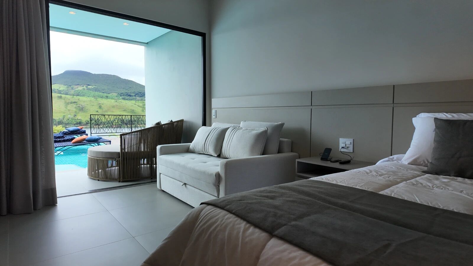 A modern hotel room with a large bed, a sofa, and a view of a swimming pool and mountains.