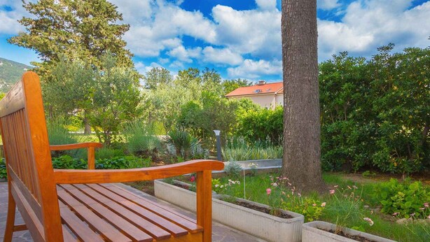 Una panchina di legno in un giardino con un vialetto in pietra, immersa nel verde e sotto un cielo sereno.