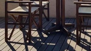 Een houten terras met tafels en stoelen, met uitzicht op een rustige zee en bergen in de verte.