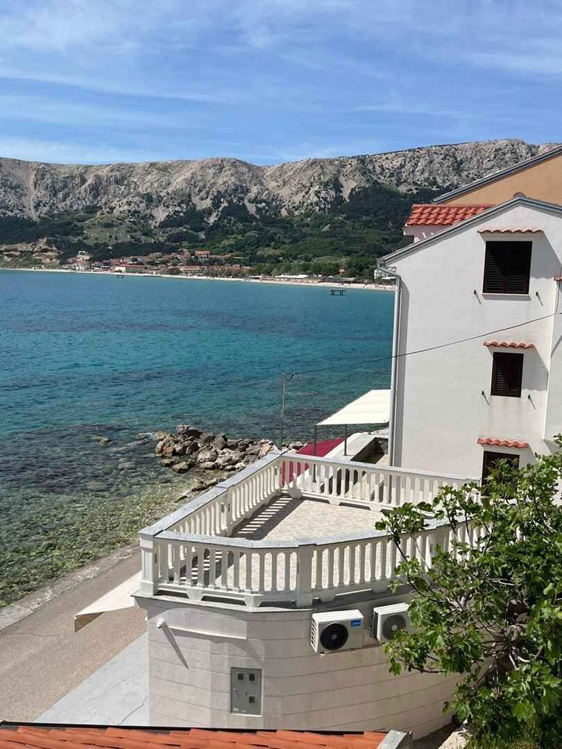 Ein Küstenhaus mit einem Balkon mit Blick auf klares blaues Wasser und einen felsigen Hügel im Hintergrund.
