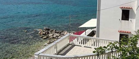 Un bâtiment situé sur la côte, doté d’un balcon donnant sur une eau d’un bleu limpide, avec une colline rocheuse en arrière-plan.