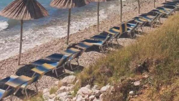 Een strand met ligbedden en rieten parasols, heuvels in de verte en een heldere blauwe zee.