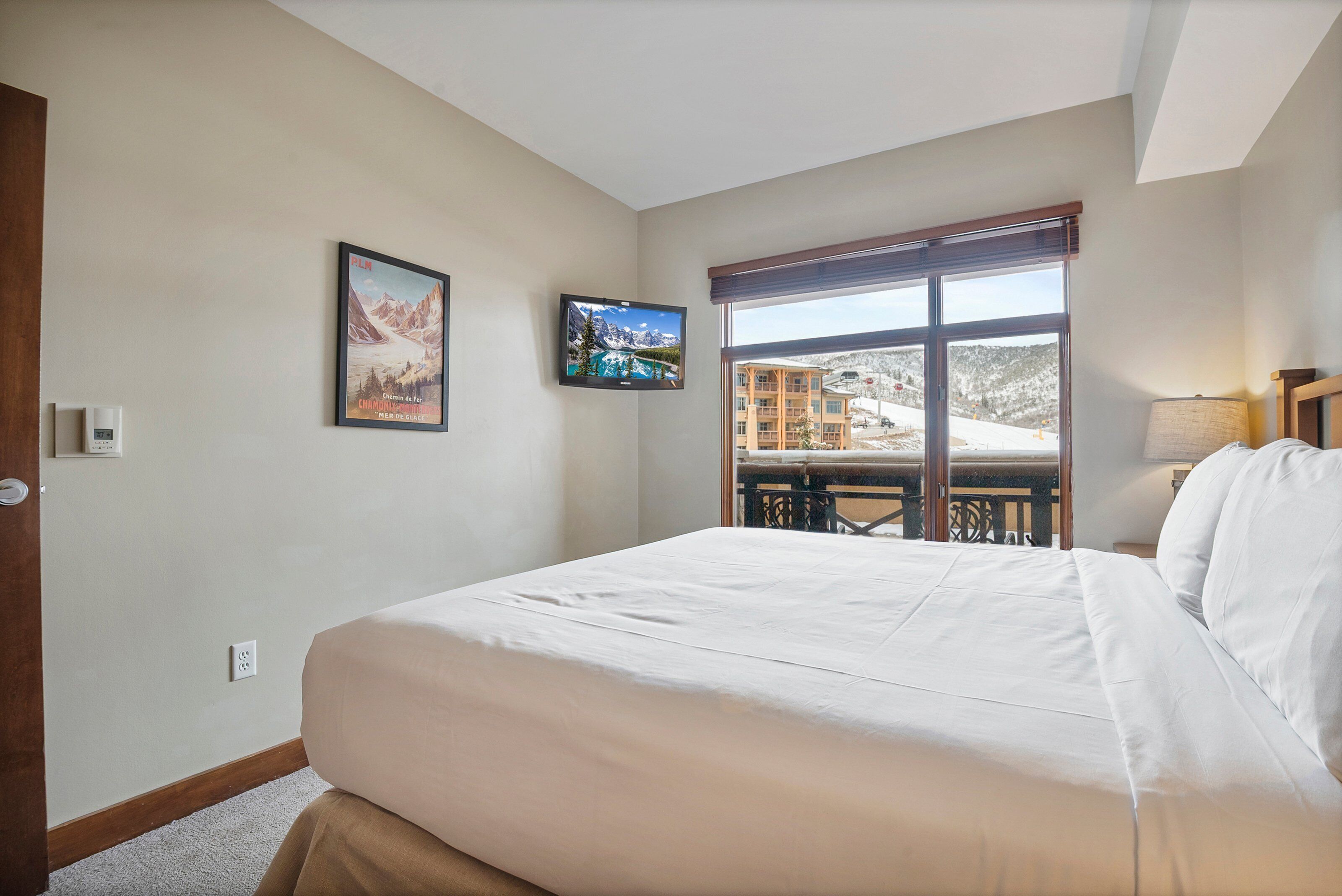 A hotel room with a large bed, a TV mounted on the wall, and a window offering a view of a building with a balcony.