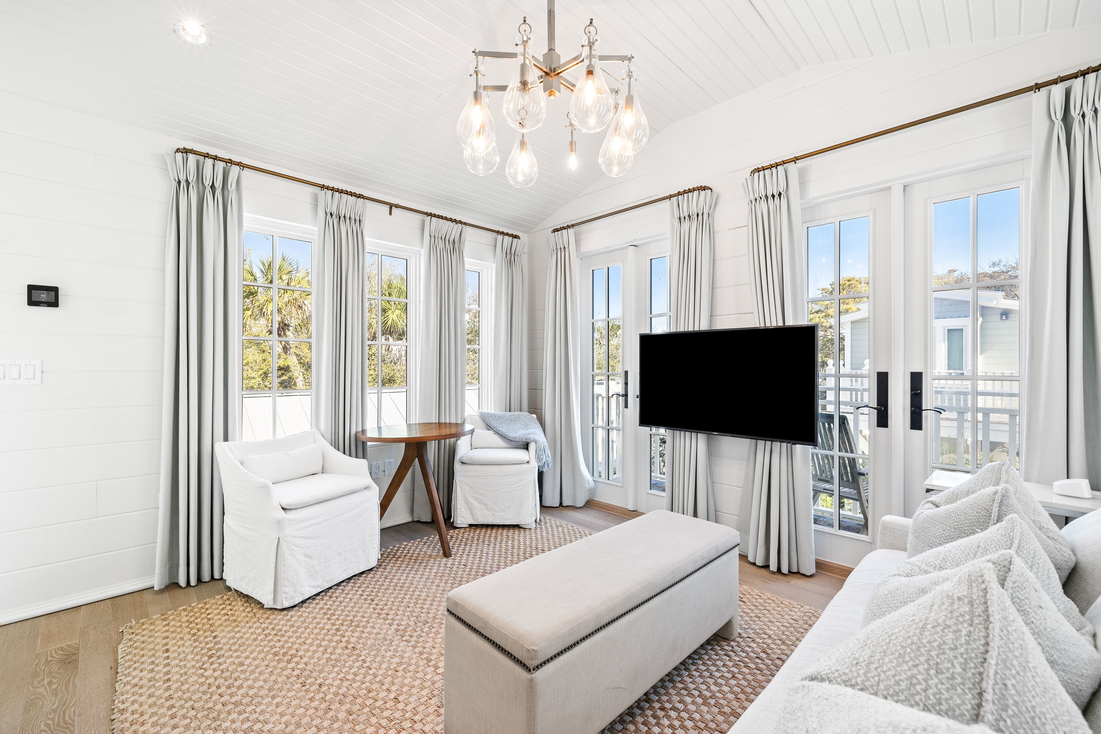 Un salón amplio con un televisor de pantalla plana grande, muebles blancos y vistas al exterior a través de amplias ventanas.