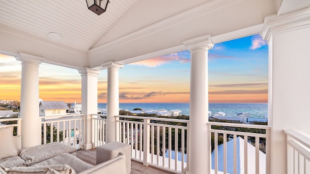Un porche cubierto con columnas blancas, asientos acolchados y vistas al océano y casas de playa.