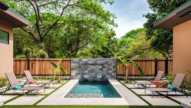 A rectangular swimming pool with a stone wall in the center, surrounded by lounge chairs and a wooden fence.