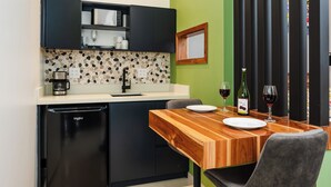 A compact kitchen with a microwave, sink, and a small refrigerator. There is a wooden dining table with chairs and a wine glass.