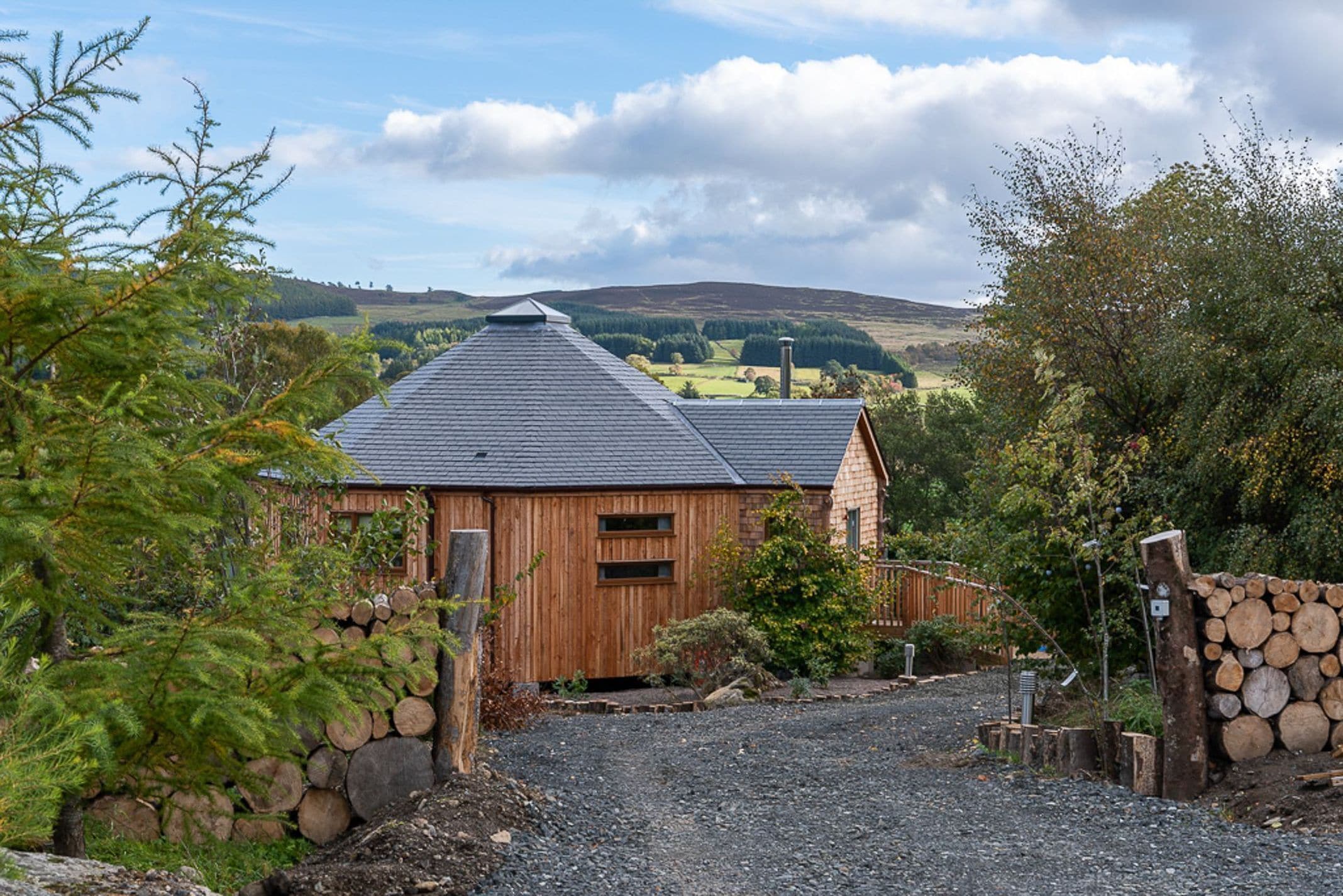 A wooden cabin with a gravel driveway and a stone wall, surrounded by trees and hills in the background.