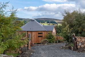 A wooden cabin with a gravel driveway and a stone wall, surrounded by trees and hills in the background. - Silver Birch Treehouse -  a treehouse that sleeps 2 guests  in 0 bedrooms (Glenshee)