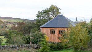 Eine Holzhütte mit geneigtem Dach, umgeben von einer Steinmauer und üppiger Vegetation.