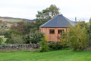 A wooden cabin with a sloped roof, surrounded by a stone wall and lush greenery.