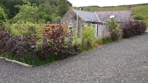 A stone cottage with a sloped roof, surrounded by a gravel driveway and lush greenery. - Smiddy Cottage -  a cottage that sleeps 4 guests  in 2 bedrooms (Glenshee)